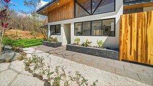 A modern two-story house with a wooden facade, large windows, and a landscaped garden with gravel and plants. - Casa Ceiba: Lush 3BR Escape Near the Ocean (Paraíso)