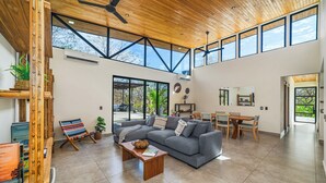 A spacious living room with a grey sofa, wooden coffee table, and a dining area with a wooden table and chairs. Large windows allow natural light and provide views of the outdoors. - Casa Ceiba: Lush 3BR Escape Near the Ocean (Paraíso)