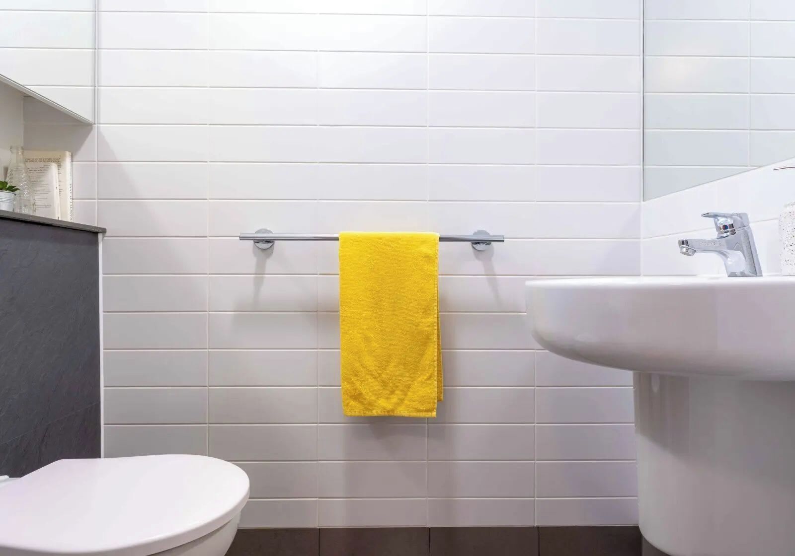 A bathroom with white tiled walls, a yellow towel hanging on a metal towel rack, a white toilet, and a white sink with a chrome faucet.