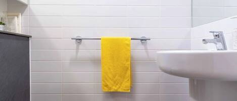 A bathroom with white tiled walls, a yellow towel hanging on a metal towel rack, a white toilet, and a white sink with a chrome faucet.