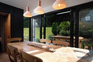 Uma área de refeições com mesa e cadeiras de madeira, janelas amplas e luminárias pendentes.