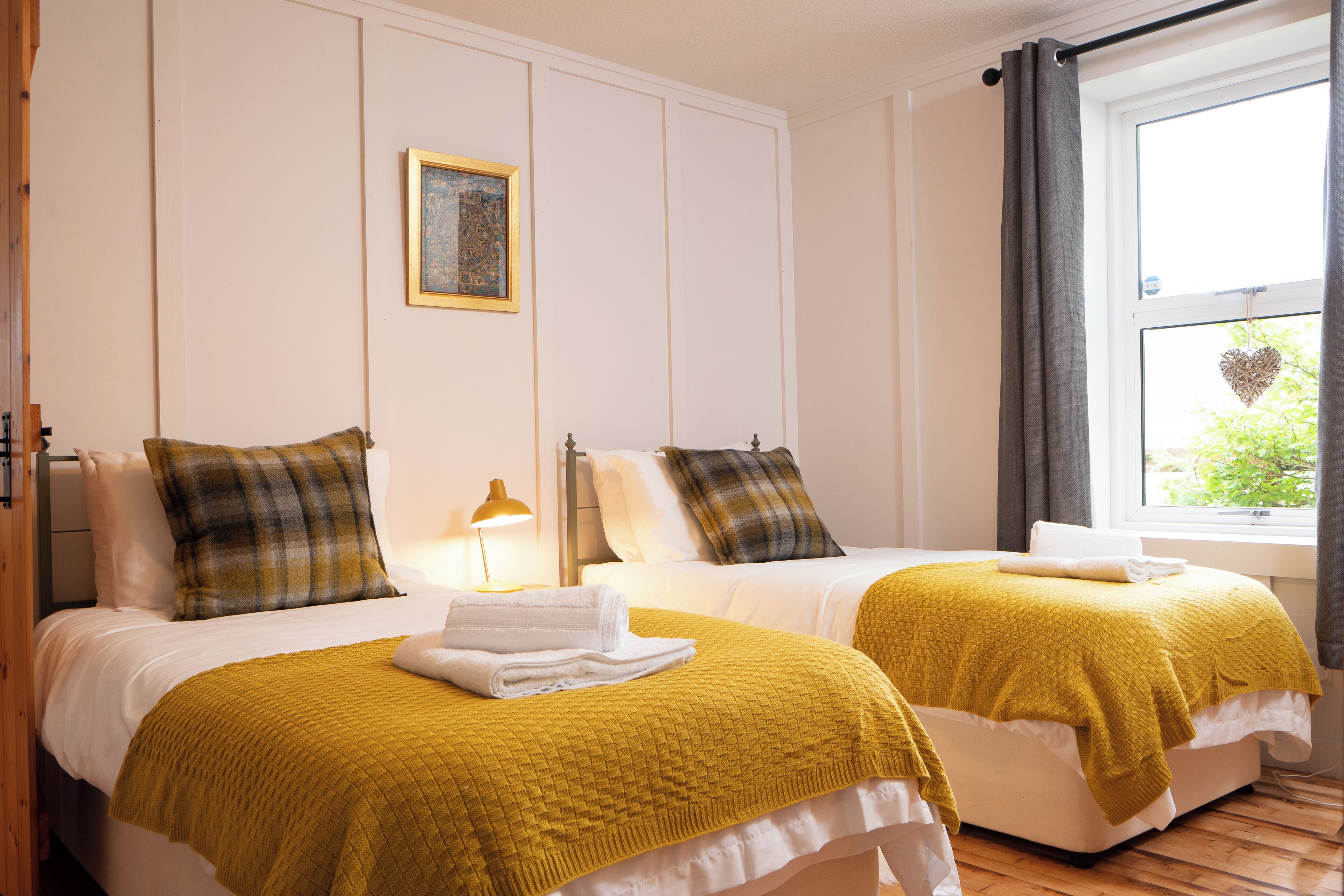 A hotel room with two beds, each with a yellow blanket and a white pillow. There is a painting on the wall and a window with a heart decoration.