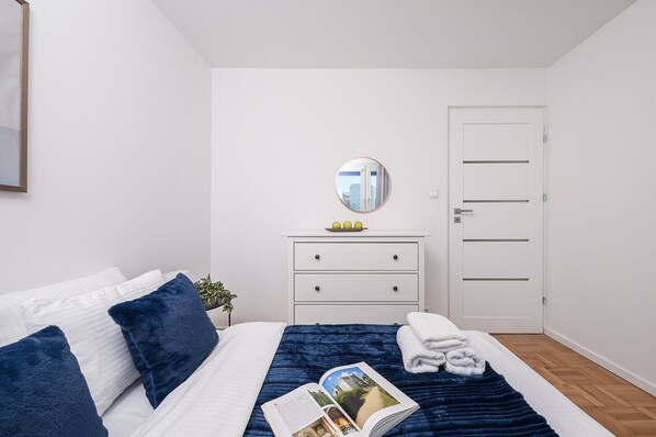 A neatly arranged bedroom with a bed, pillows, a bedside table with a mirror, a door, and a small plant. - Lumina apart with balcony in Warsaw (Warsaw)