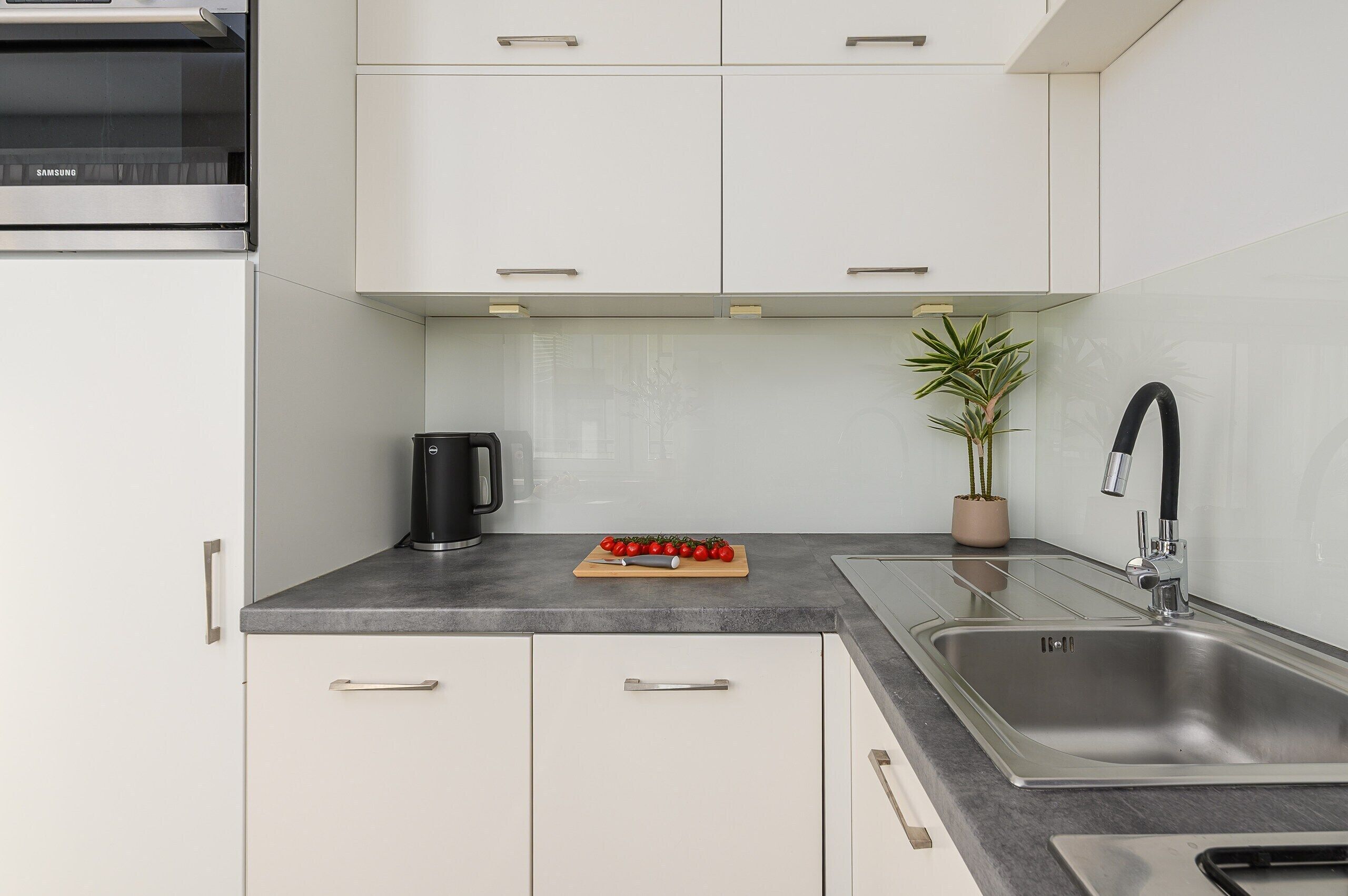 A modern kitchen with white cabinets, a stainless steel sink, a black coffee maker, and a wooden cutting board with tomatoes.