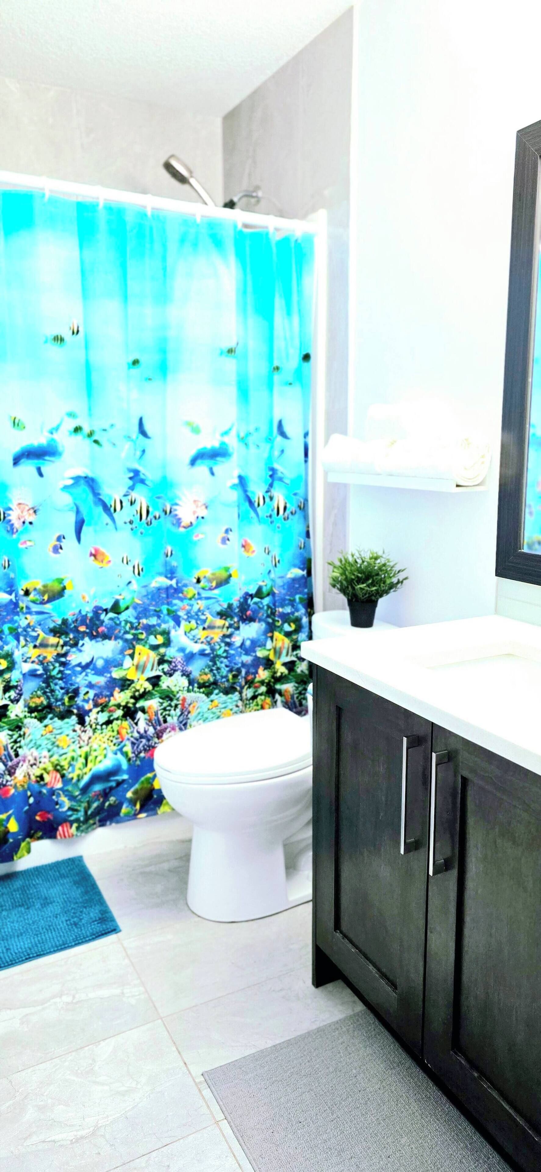 Une salle de bain avec des toilettes blanches, des meubles en bois foncé, un plan de travail blanc, un rideau de douche orné d’un motif représentant un paysage marin bleu, et une petite plante en pot posée sur le plan de travail.