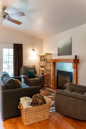 A living room with a fireplace, a ceiling fan, a bookshelf, and a window with curtains.