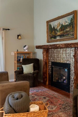 Un salon chaleureux avec une cheminée, un tableau, une bibliothèque et un fauteuil confortable.