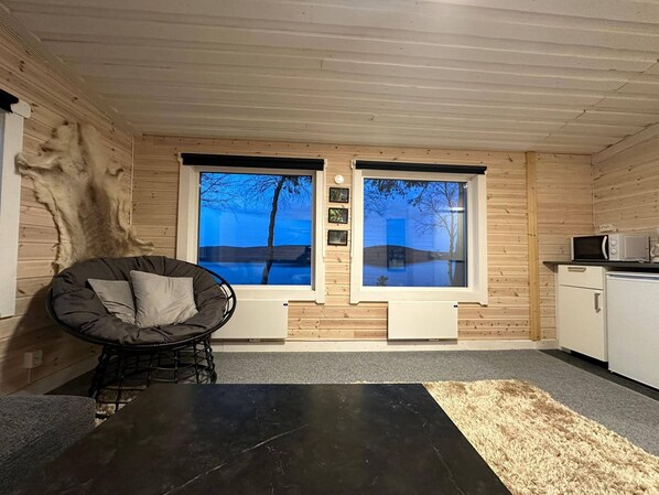 A cozy cabin interior with a large window, a chair, a small kitchen area, and a black floor.