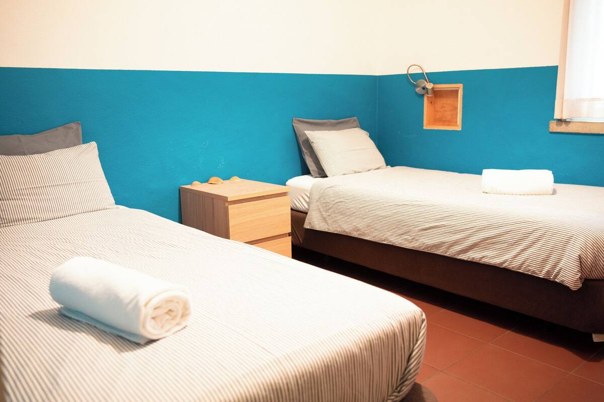 A hotel room with two beds, each with a pillow and a rolled towel. There is a wooden nightstand with a lamp and a small wooden box.