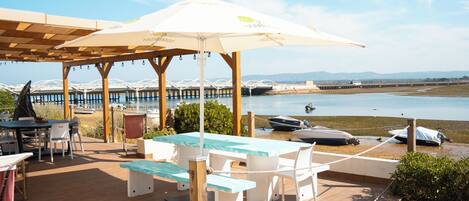 A wooden deck with a table and chairs, a covered area with an umbrella, and boats in the background.