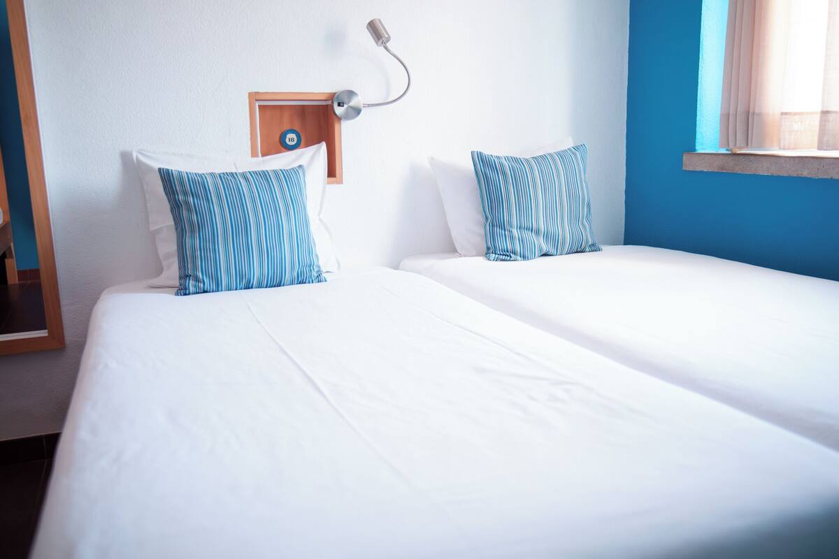 A hotel room with two beds, a wooden headboard, a bedside lamp, and a window with curtains.