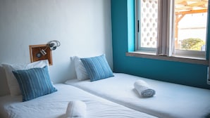 A hotel room with two beds, each with white linens and blue striped pillows. There is a window with a view of the outside, and a wooden headboard with a light fixture. - Wax Hostel & Apts (Faro)