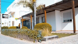 A white building with a sign for a hostel and an ATM. There are neatly trimmed bushes and a palm tree. - Wax Hostel & Apts (Faro)