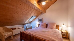 Una camera accogliente con soffitto spiovente, un letto di legno, una poltrona in vimini, un comodino con una lampada e un quadro incorniciato appeso al muro.