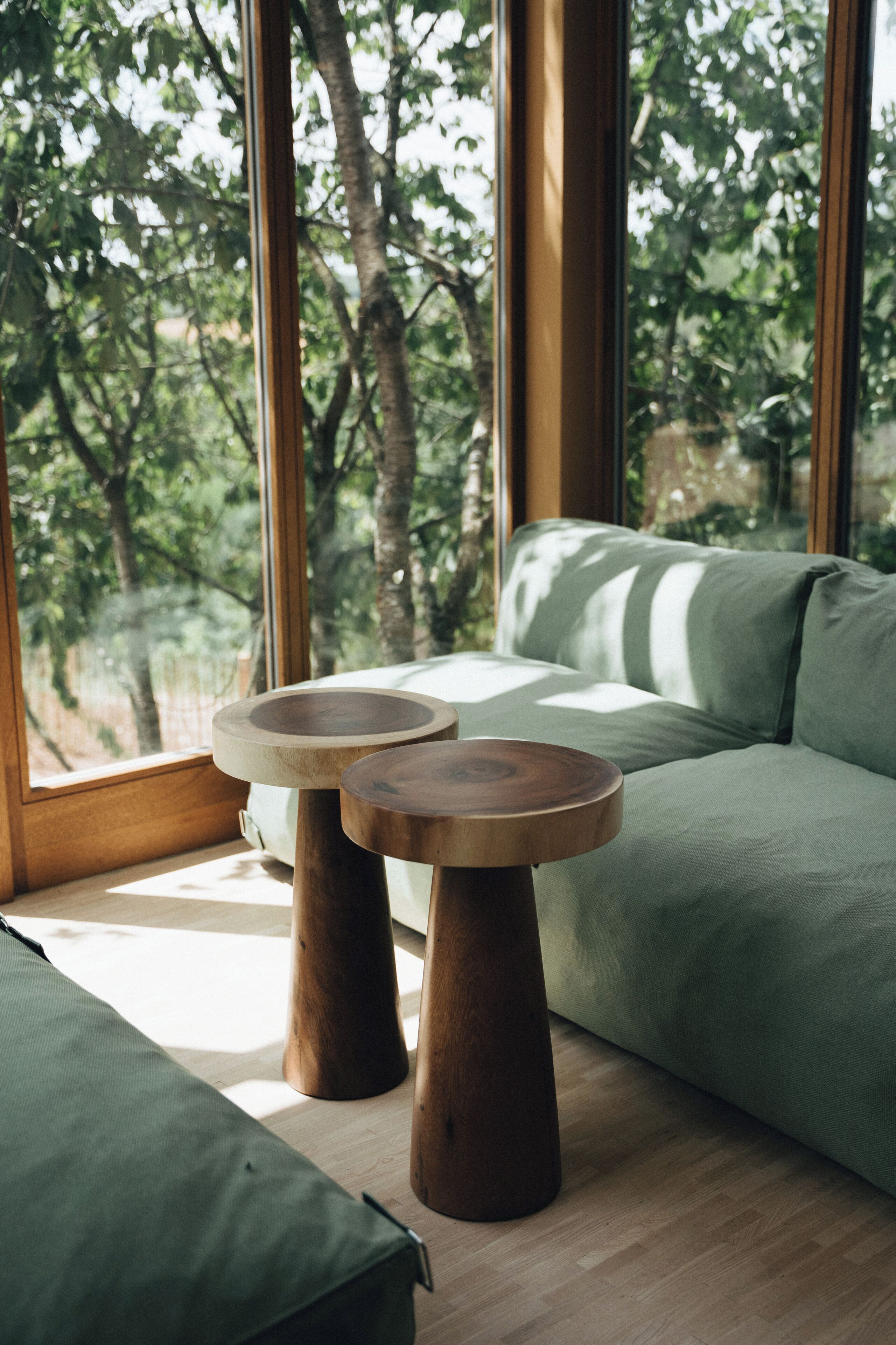 Een kamer met een groene bank, twee houten tafels en grote ramen met uitzicht op bomen.