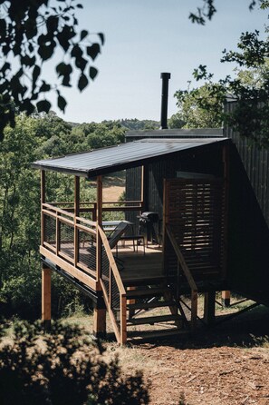 Una terraza de madera con techo metálico, rodeada de árboles y vista a zona boscosa.