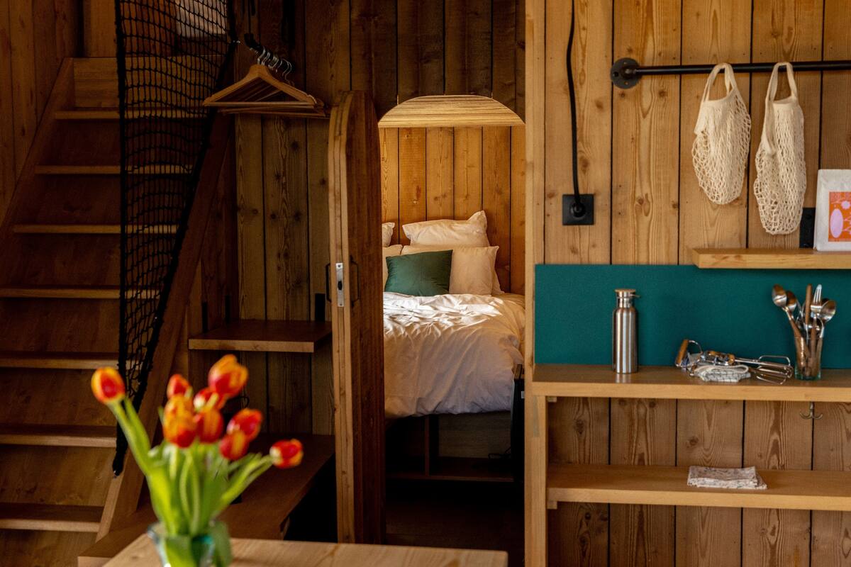 A wooden interior with a bed, a hanging light, a shelf with kitchen utensils, and a staircase.