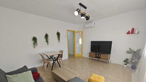 Una sala de estar moderna con un sofá gris, una mesa de centro de madera, un mueble para TV y un pequeño reposapiés amarillo. Hay una mesa de comedor con sillas y plantas colgantes en la pared.