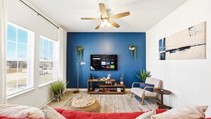 A living room with a red sofa, a ceiling fan, a TV mounted on the wall, and a window with blinds. - Unit 101 Family-friendly stay San Antonio 2BR for 6 (San Antonio)