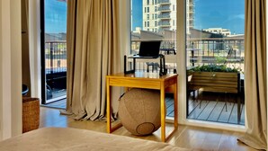 Moderni hotellihuone, jossa on suuri ikkuna, puinen työpöytä kannettavalla tietokoneella, mukava tuoli ja näkymä kaupunkimaisemaan.