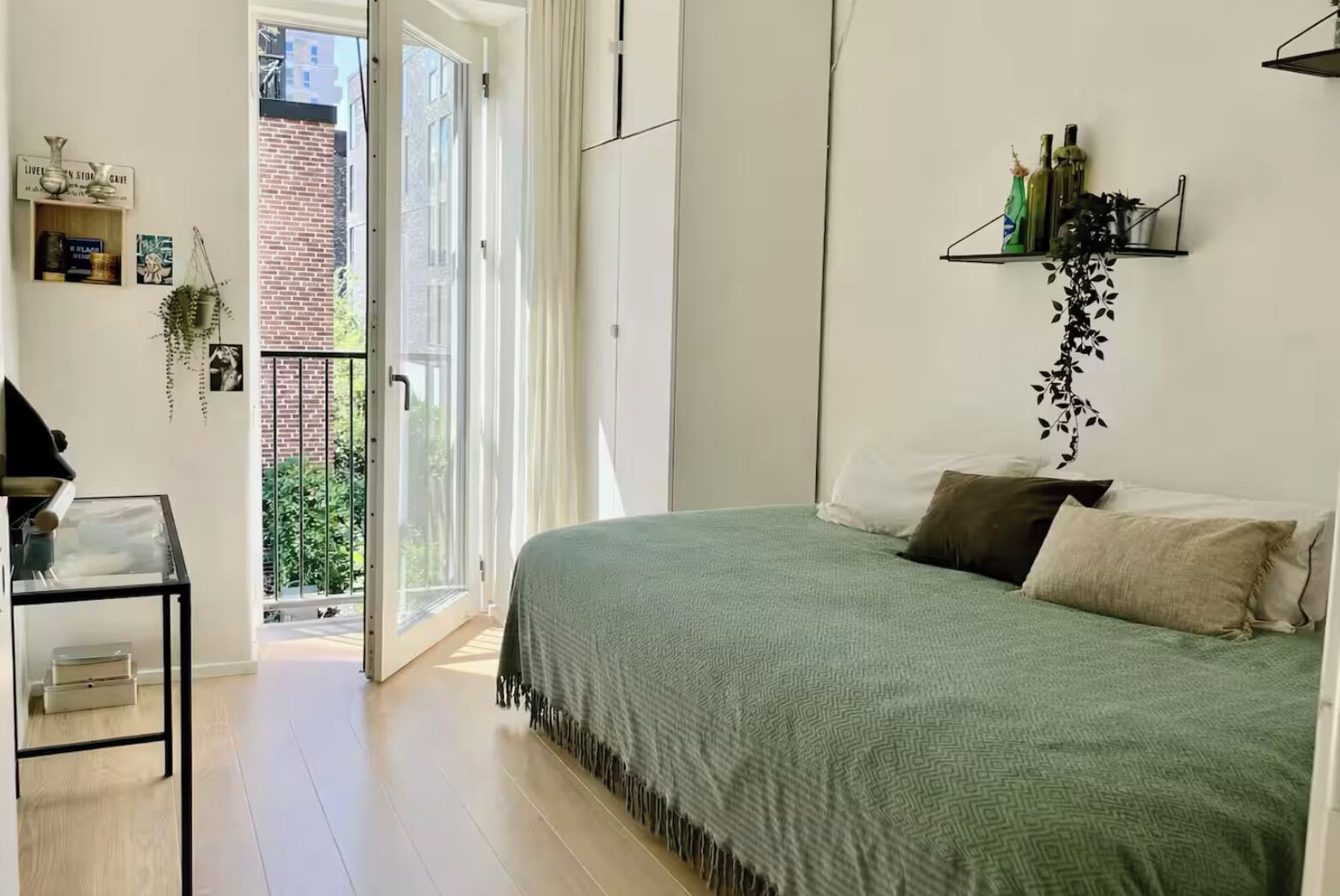 Een slaapkamer met een bed, een klein tafeltje, een plank met flessen en uitzicht op een bakstenen gebouw door een deur.