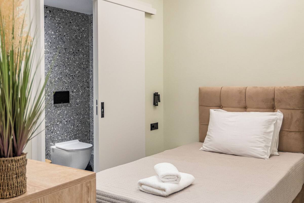 A bedroom with a bed, a bedside table, a wooden nightstand, a wall-mounted light, and a door leading to a bathroom.