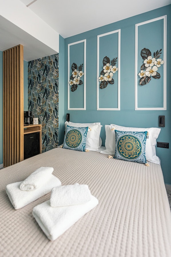 A hotel room with a bed, two pillows, a wall with floral decorations, a wooden panel, and a coffee machine. - Ernest Suites Psiri (Athens)
