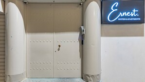 Hotellin sisäänkäynti, jossa on vaaleanbeige väriin maalattu kaksoisovi, sen yläpuolella valvontakamera ja kyltti, jossa lukee ”Ernest Suites & Apartments”.