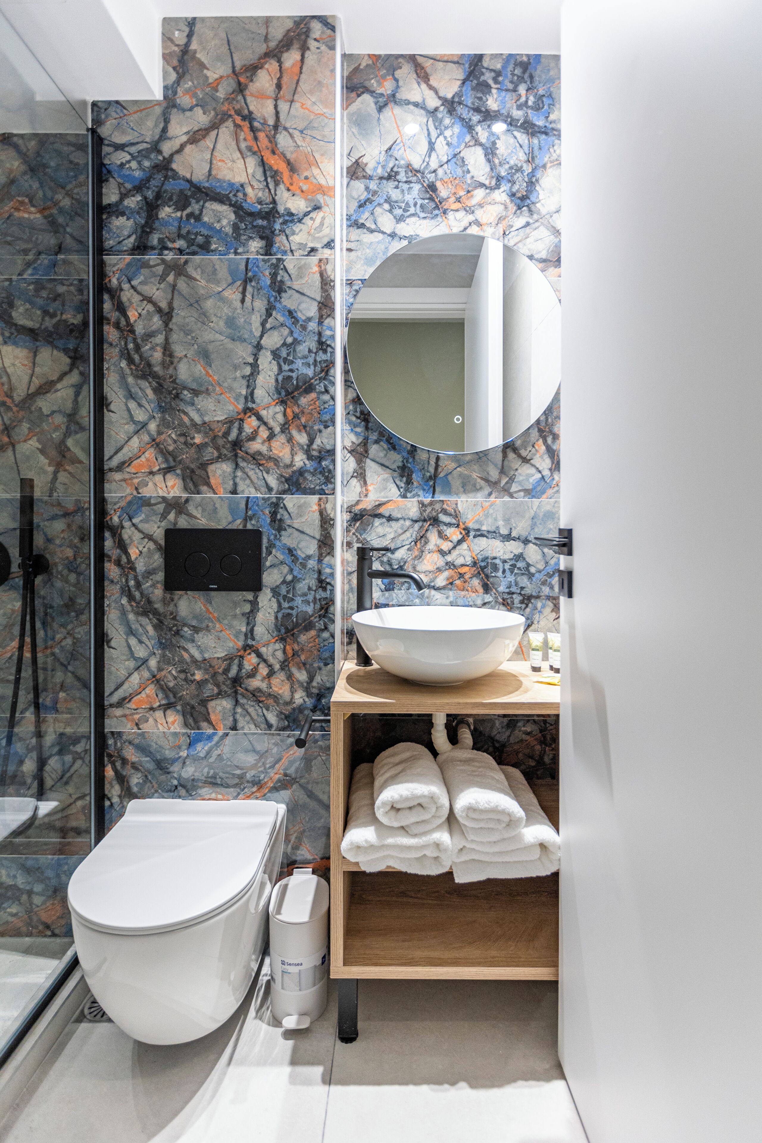 A modern bathroom with a glass-enclosed shower, a white toilet, a marble backsplash, a round mirror, and a wooden shelf with towels.
