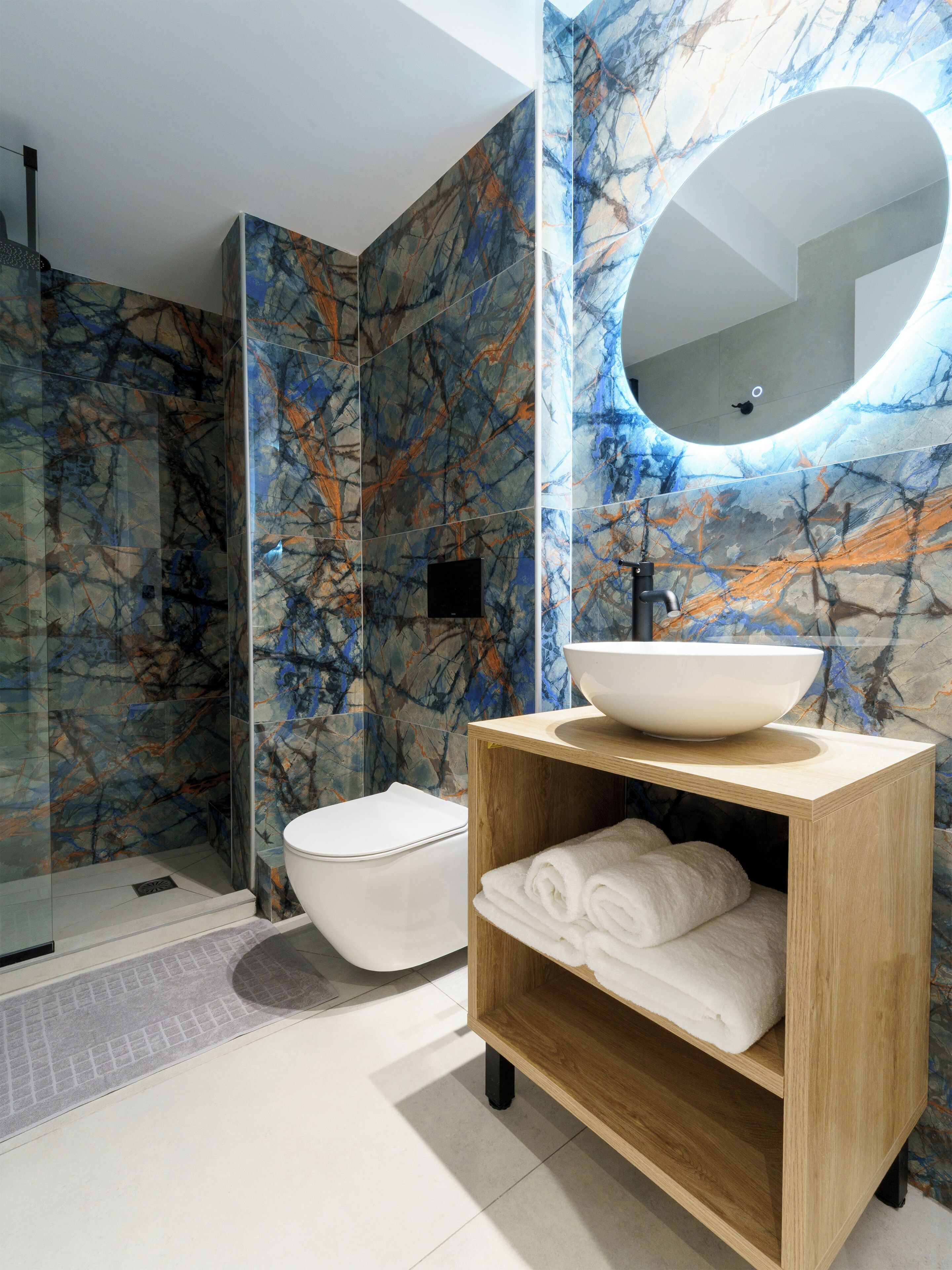 A modern bathroom with a marble wall, a round mirror, a white sink, and a wooden shelf with towels.