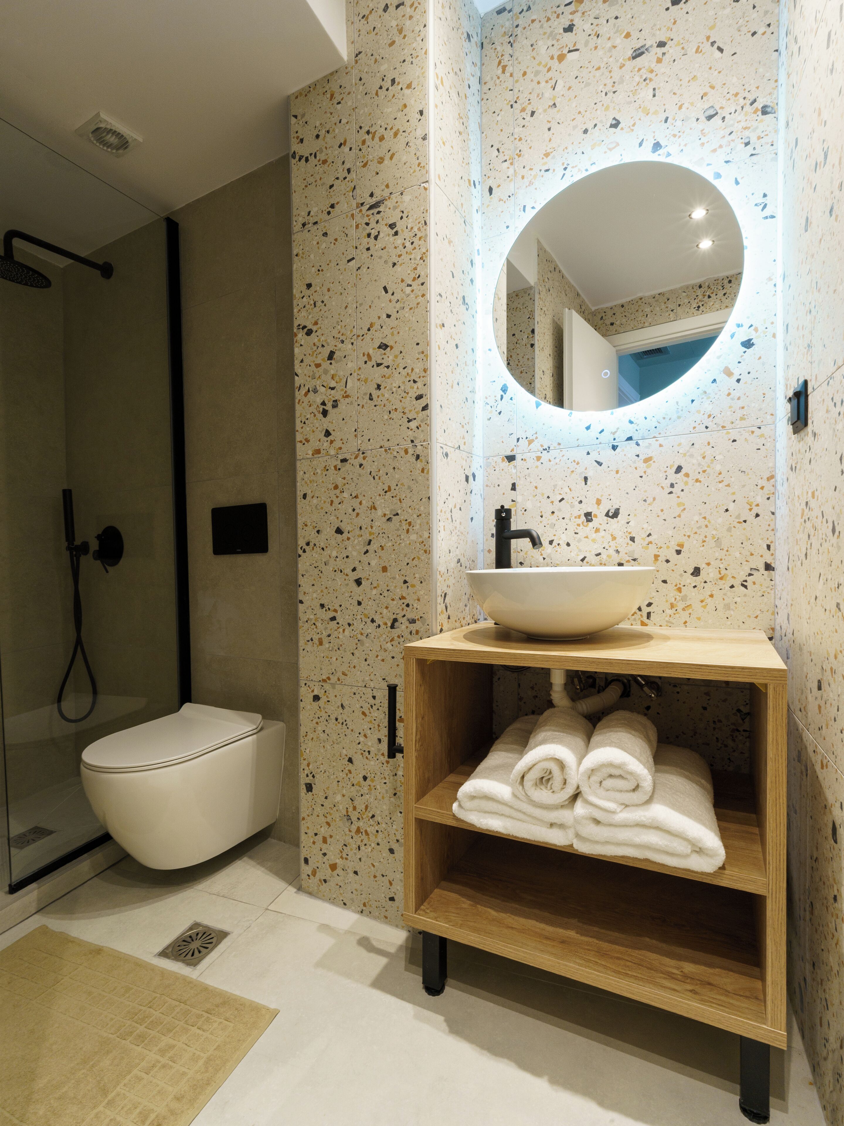 A modern bathroom with a freestanding toilet, a built-in sink with a wooden vanity, a shower enclosure, and a mirror with a circular frame.