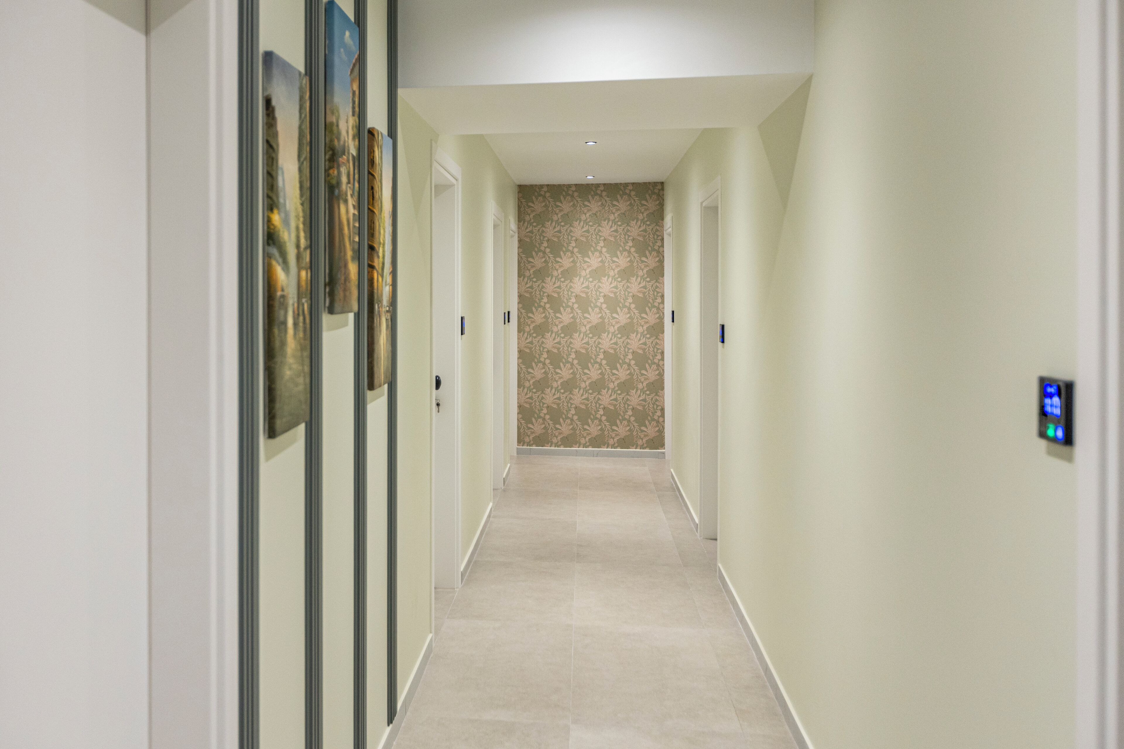 A hallway with a light-colored tiled floor, a floral wallpaper wall, and framed artwork on the left.