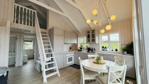 A spacious kitchen with white cabinets, a central island, and a dining area with white chairs. A white ladder is leaning against the wall, and there are multiple hanging light fixtures.