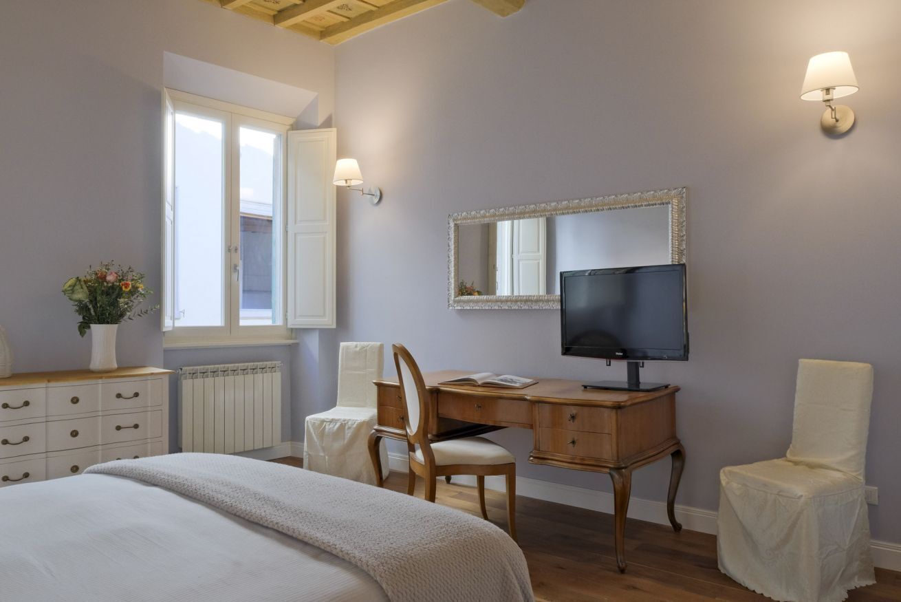 Un dormitorio con cama, escritorio, silla, televisor, espejo, radiador y ventana.
