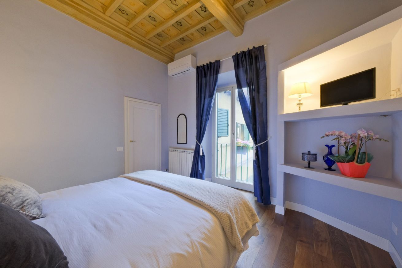 A bedroom with a bed, a TV, a shelf with decorative items, and a window with curtains.
