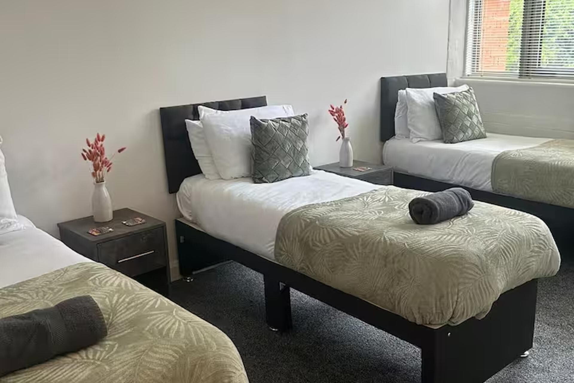 A hotel room with three single beds, each with a dark headboard, white bedding, and a grey throw blanket. There are bedside tables with lamps and a vase with flowers.