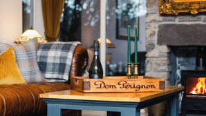Uma sala de estar aconchegante com lareira, uma caixa de madeira com 'Dom Perignon' escrito, uma garrafa e duas velas verdes sobre uma mesa.
