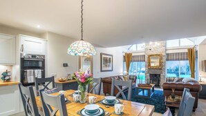 Uma cozinha moderna com fornos duplos, sala de jantar com mesa de madeira posta para quatro e sala de estar com lareira e vista para o exterior.