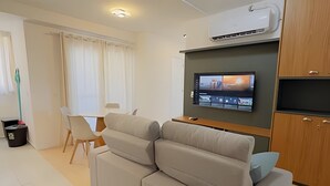 Una sala moderna con un sofá gris, una mesa de comedor con sillas, un televisor montado en la pared y un equipo de aire acondicionado sobre el sofá.