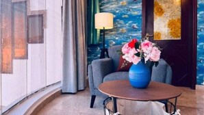 A room with a round table, a blue vase with flowers, a grey armchair, a lamp, and a door with a golden pattern.