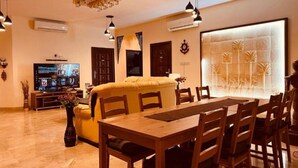 A spacious living area with a large sofa, dining table, and chairs. There is a flat-screen TV mounted on the wall and a decorative wall panel.