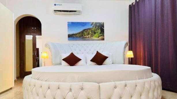 A round bed with a tufted headboard and two pillows, a wall-mounted air conditioner, and a framed picture of a landscape.