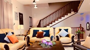 A spacious living room with two beige sofas, a coffee table with a floral centerpiece, and a staircase in the background.