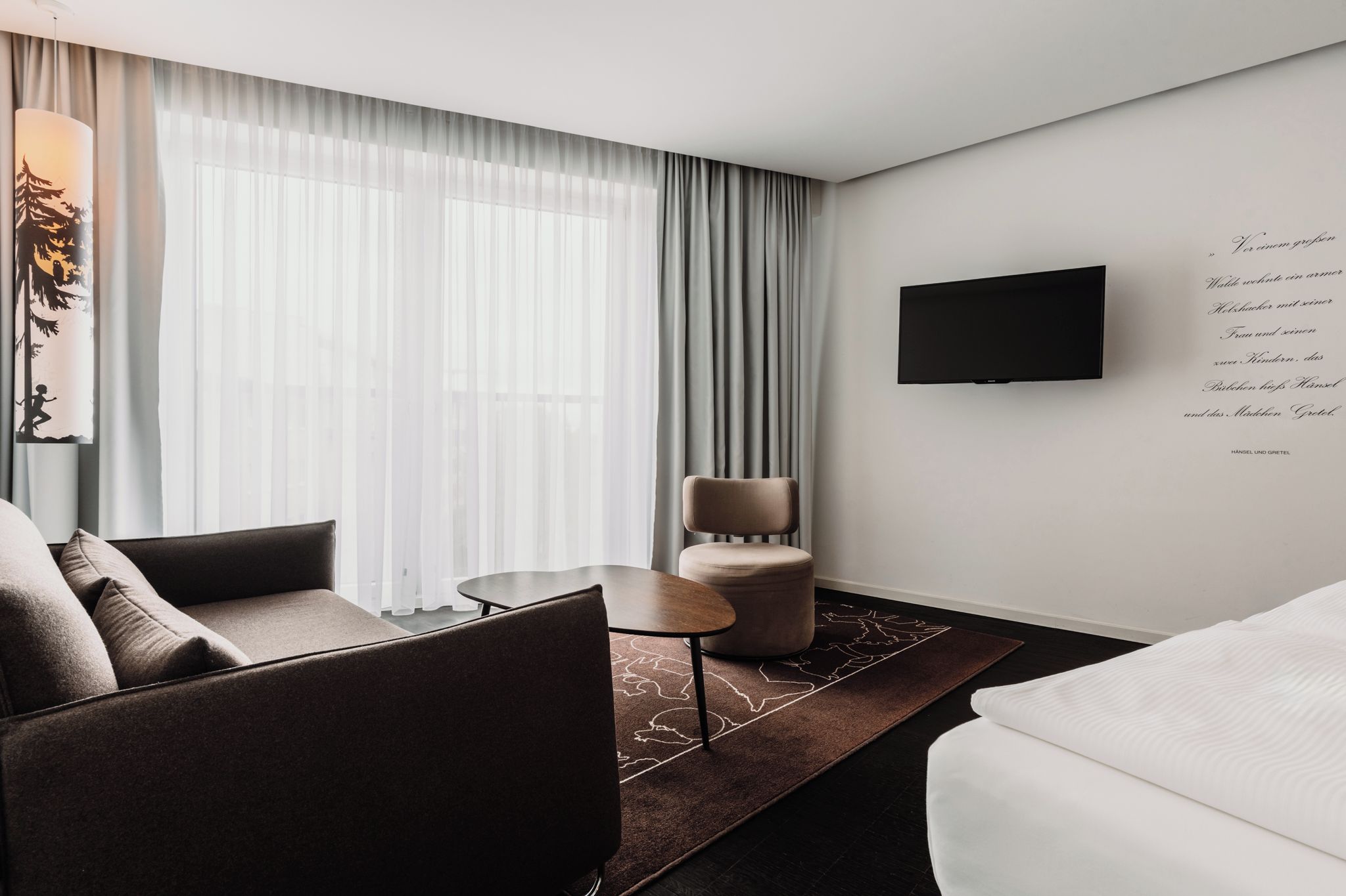 A modern hotel room with a sofa, a chair, a small table, a TV mounted on the wall, and a large window with sheer curtains.