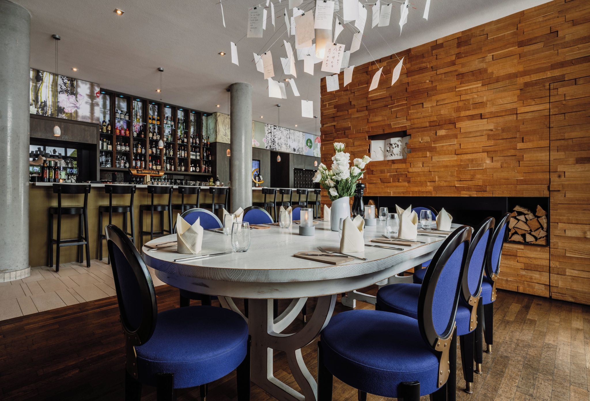 A restaurant with a round table set for dining, bar area with various bottles, and a wooden wall.