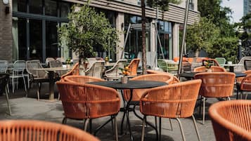 An outdoor seating area with wicker chairs and tables, a small tree, and a building with large windows in the background.