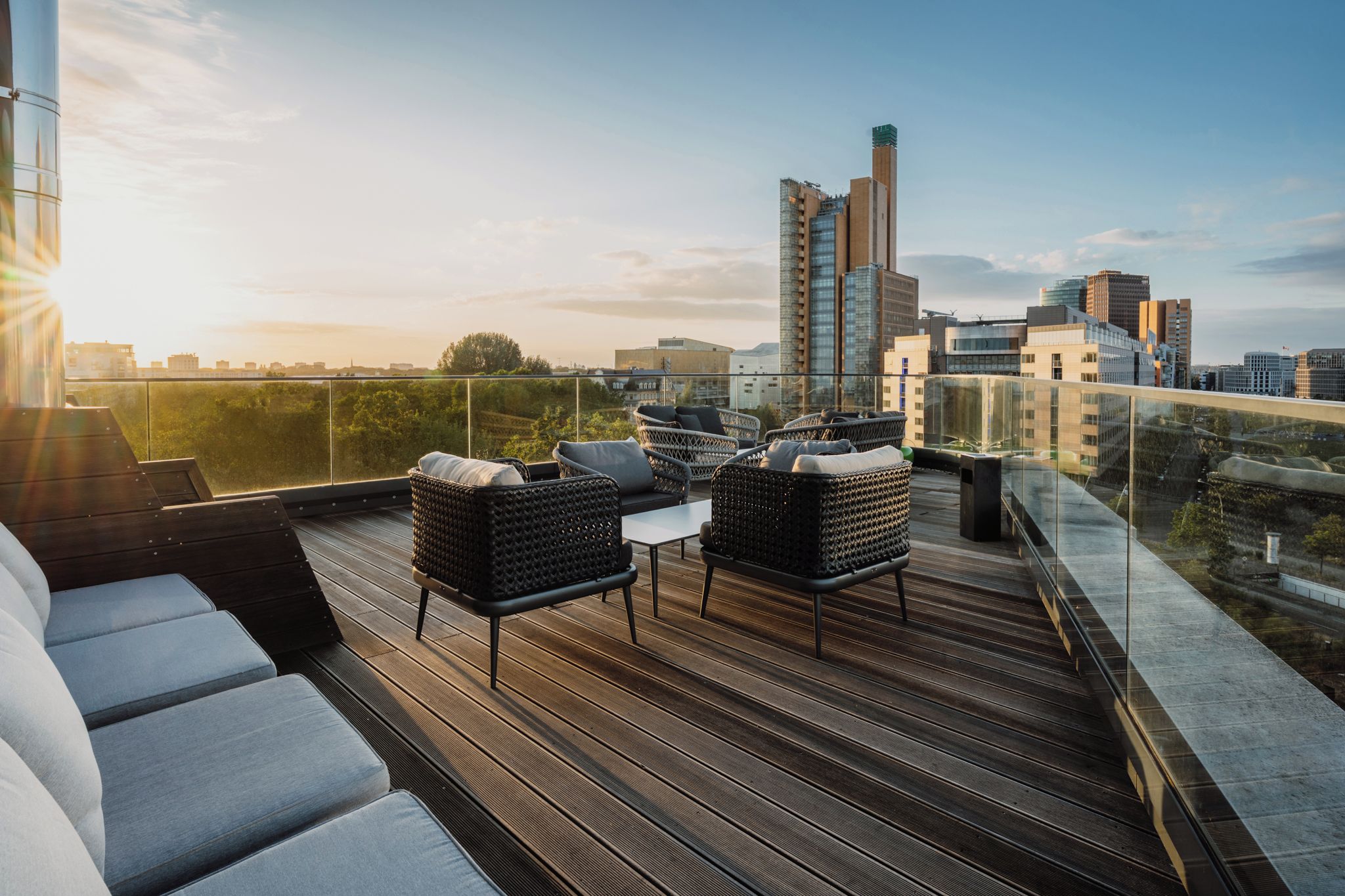 A rooftop terrace with wicker furniture, a glass balustrade, and a city skyline view.