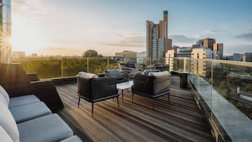 A rooftop terrace with wicker furniture, a glass balustrade, and a city skyline view.
