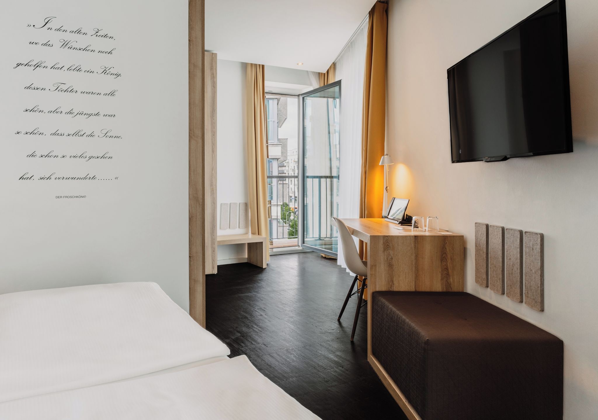 Habitación de hotel con cama, escritorio con ordenador portátil, televisor montado en la pared y vista a la ciudad por la ventana.
