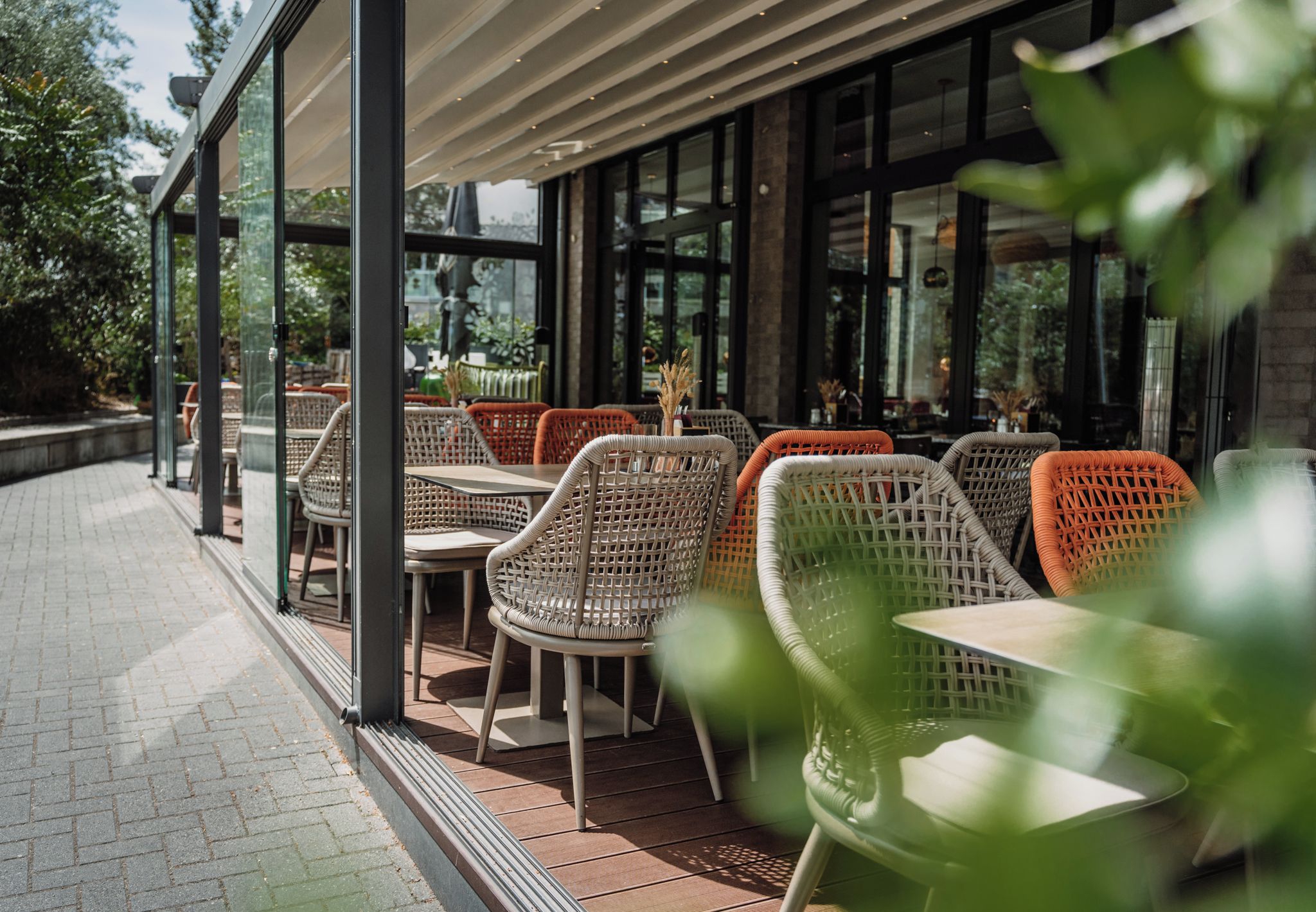 An outdoor cafe with wicker chairs and tables, large windows, and a brick pathway.